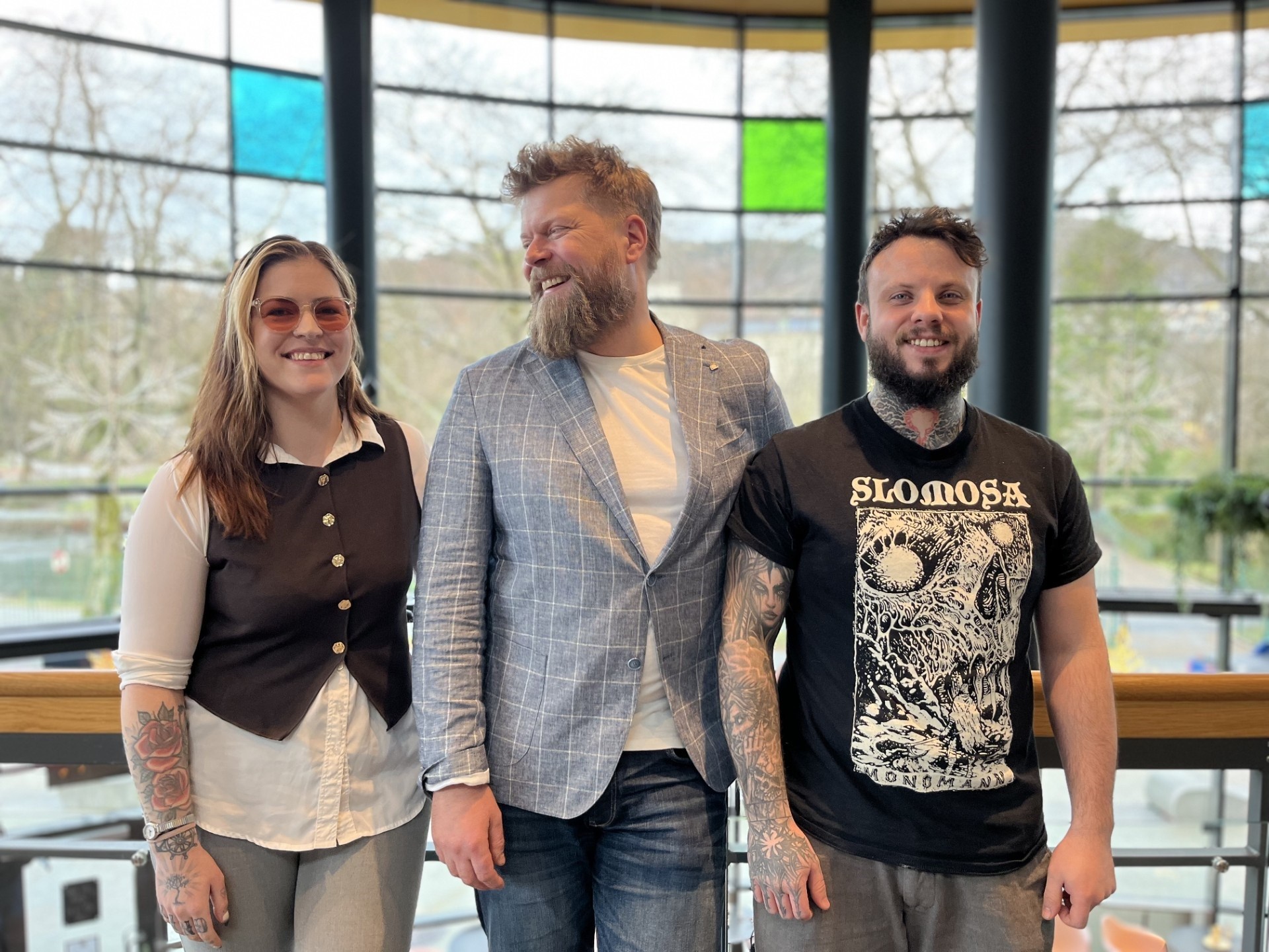 Three people stand together in a brightly lit room with large windows, smiling and posing for the camera.