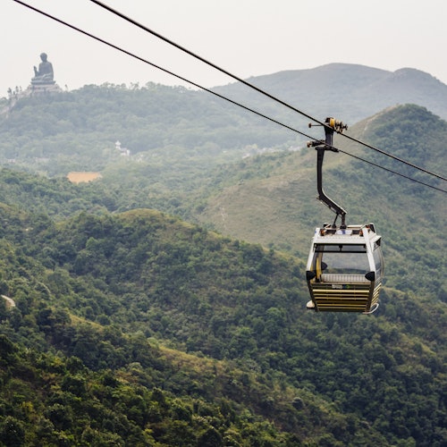 Una funivia vuota sospesa sopra un lussureggiante paesaggio montuoso con una grande statua di Buddha visibile in lontananza.