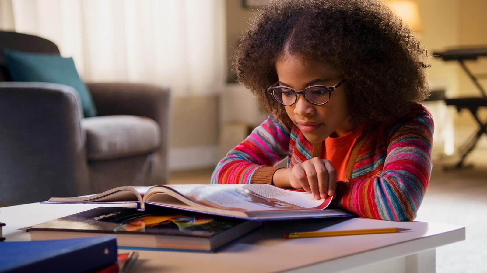 A girl wearing glasses and a colorful sweater reads a book at a white table, with other books and a pencil nearby in a living room setting.