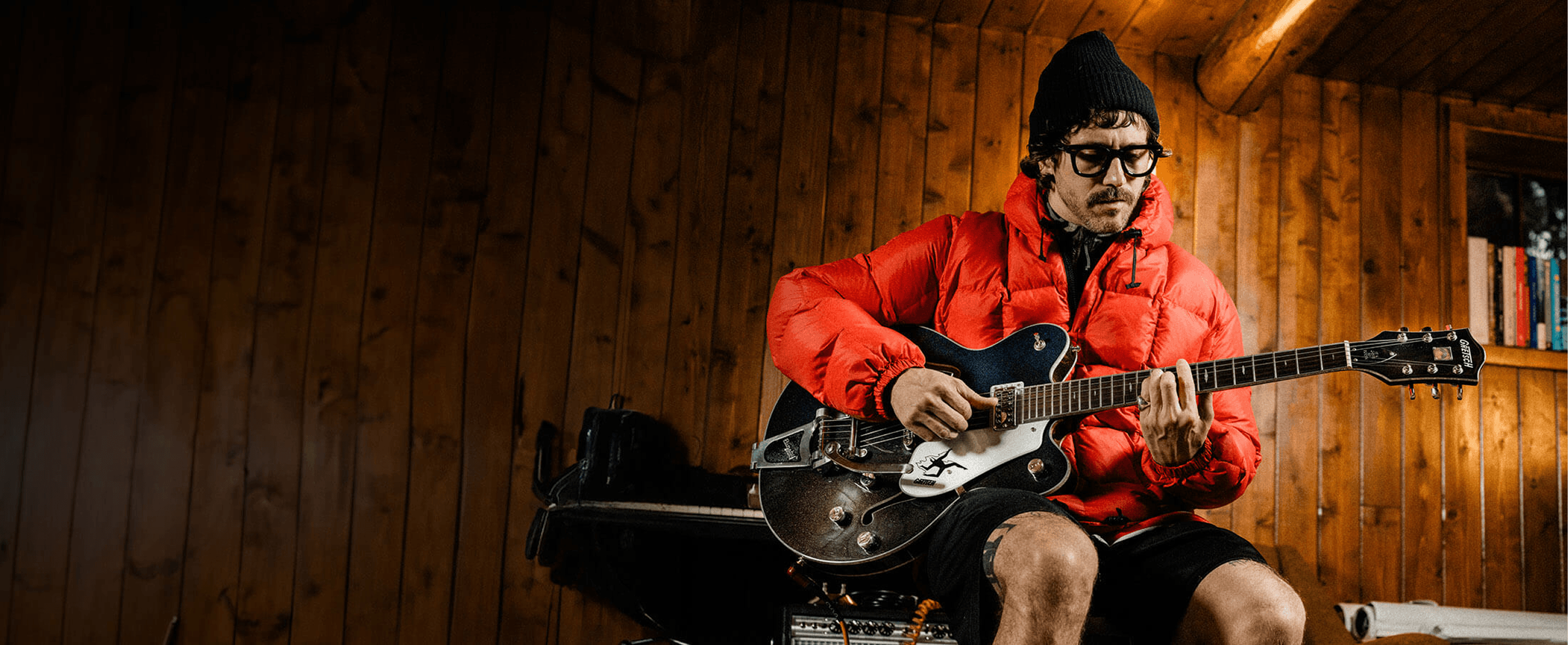 Portugal The Man's John Gourley in a red puffer jacket playing a Gretsch electric guitar inside a warm wood-paneled cabin.