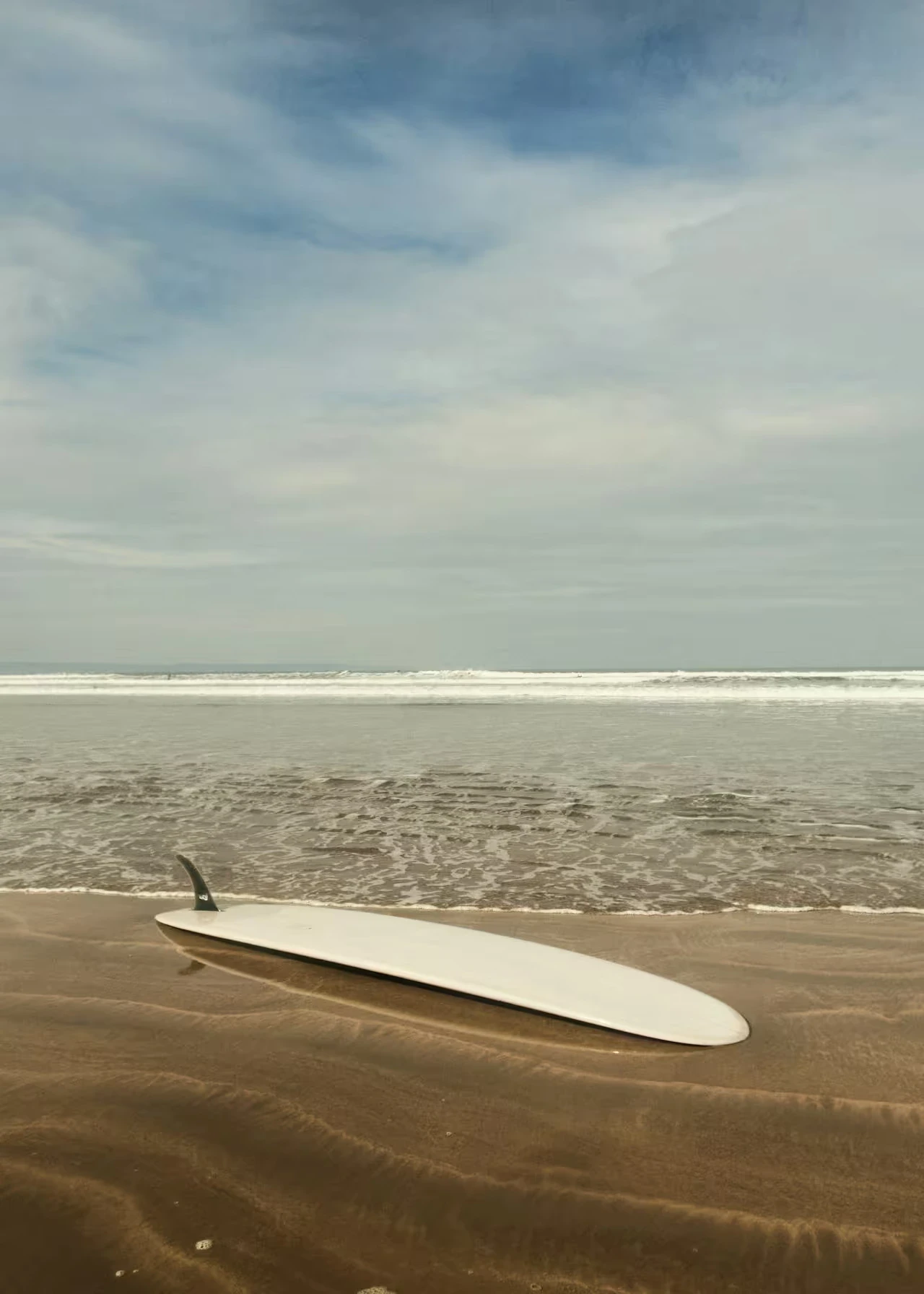 A white surfboard rests fin-up on wet sand at the shoreline with ocean waves in the background.