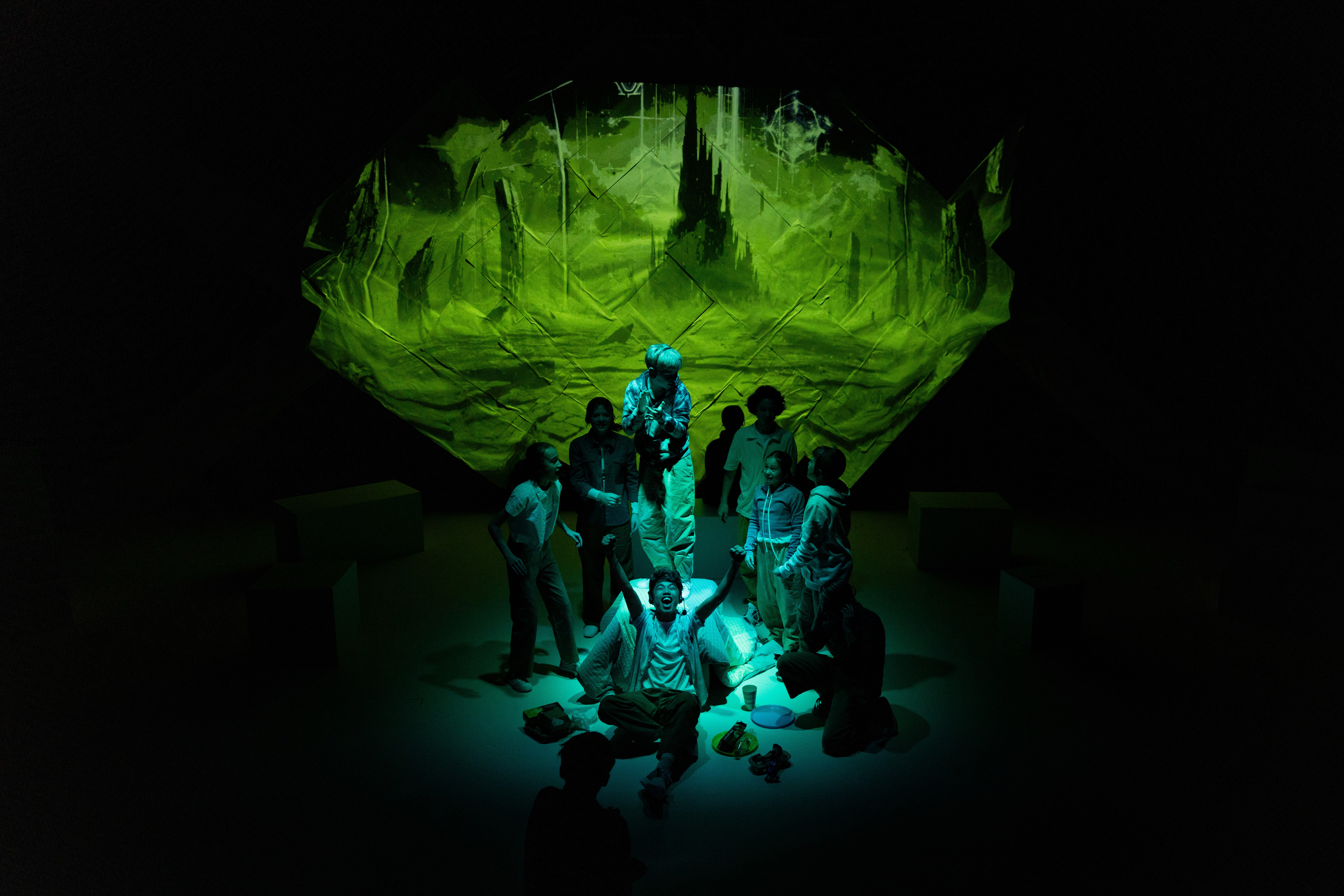A group of young performers gather in a circle around a central figure, looking up at a green illuminated projection resembling a dark, towering sci-fi landscape.