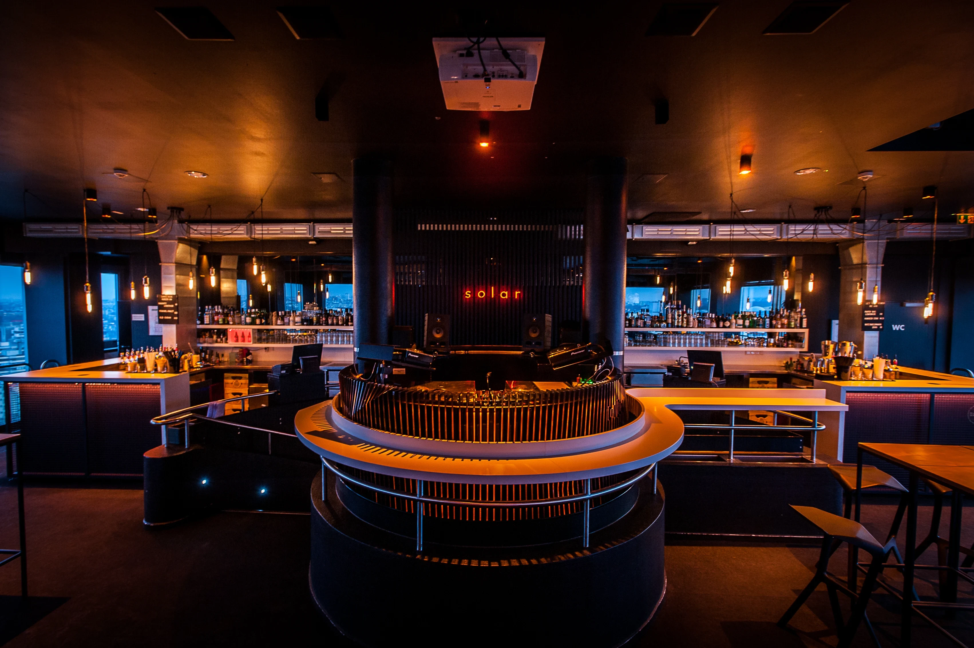 Panoramic view of SOLAR SKY LOUNGE with a circular central bar with brass railing, the red solar neon above, long illuminated counters and dark ceilings with hanging bulbs.