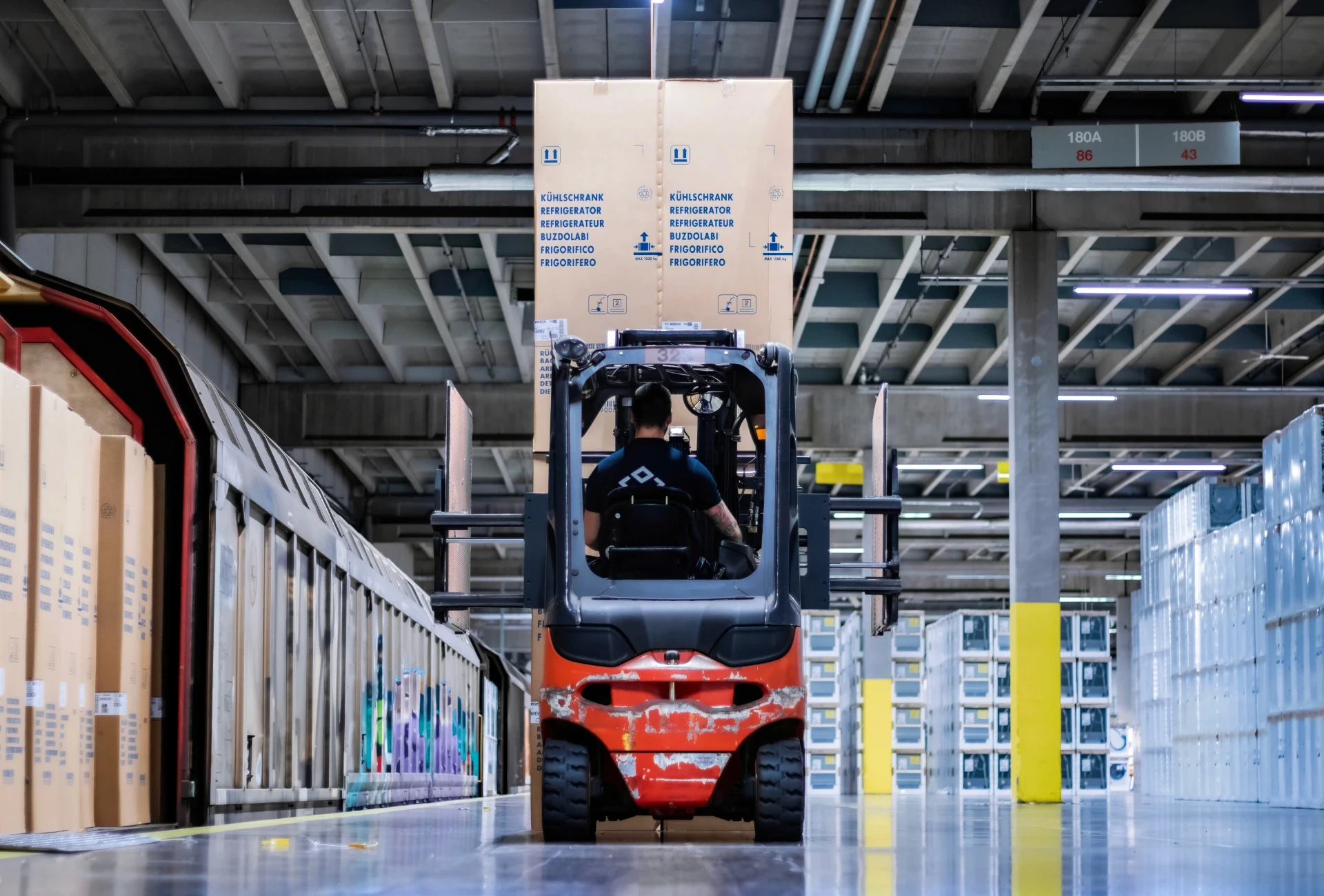 En gaffeltruck lyfter en stor låda i ett lager. En person kör trucken.