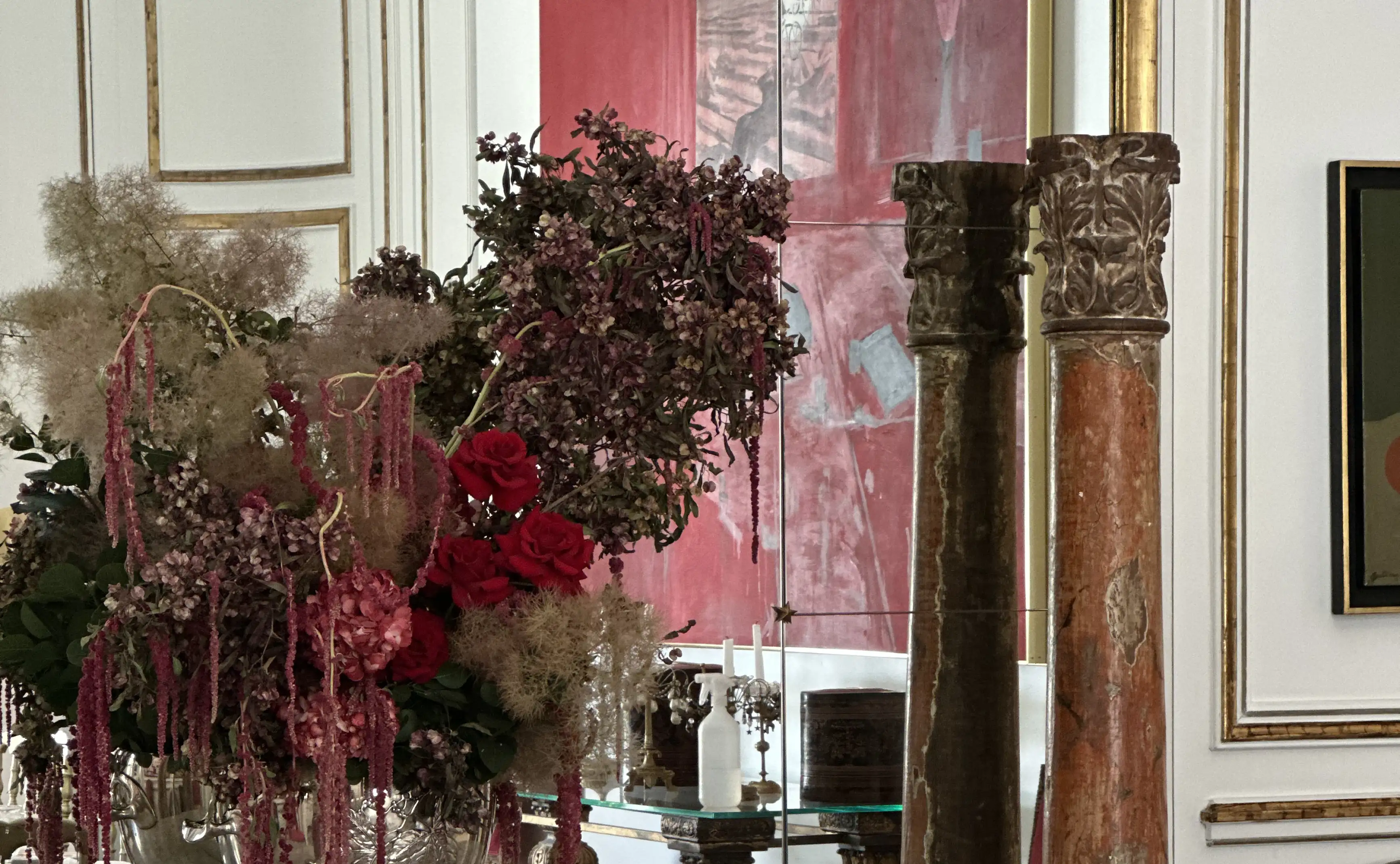 Arreglo floral exuberante con rosas rojas, hortensias y ramas colgantes en tonos púrpura y beige, dispuesto en una base plateada frente a un espejo con columnas antiguas y un fondo artístico en color rojo, dentro de un salón clásico con molduras doradas.