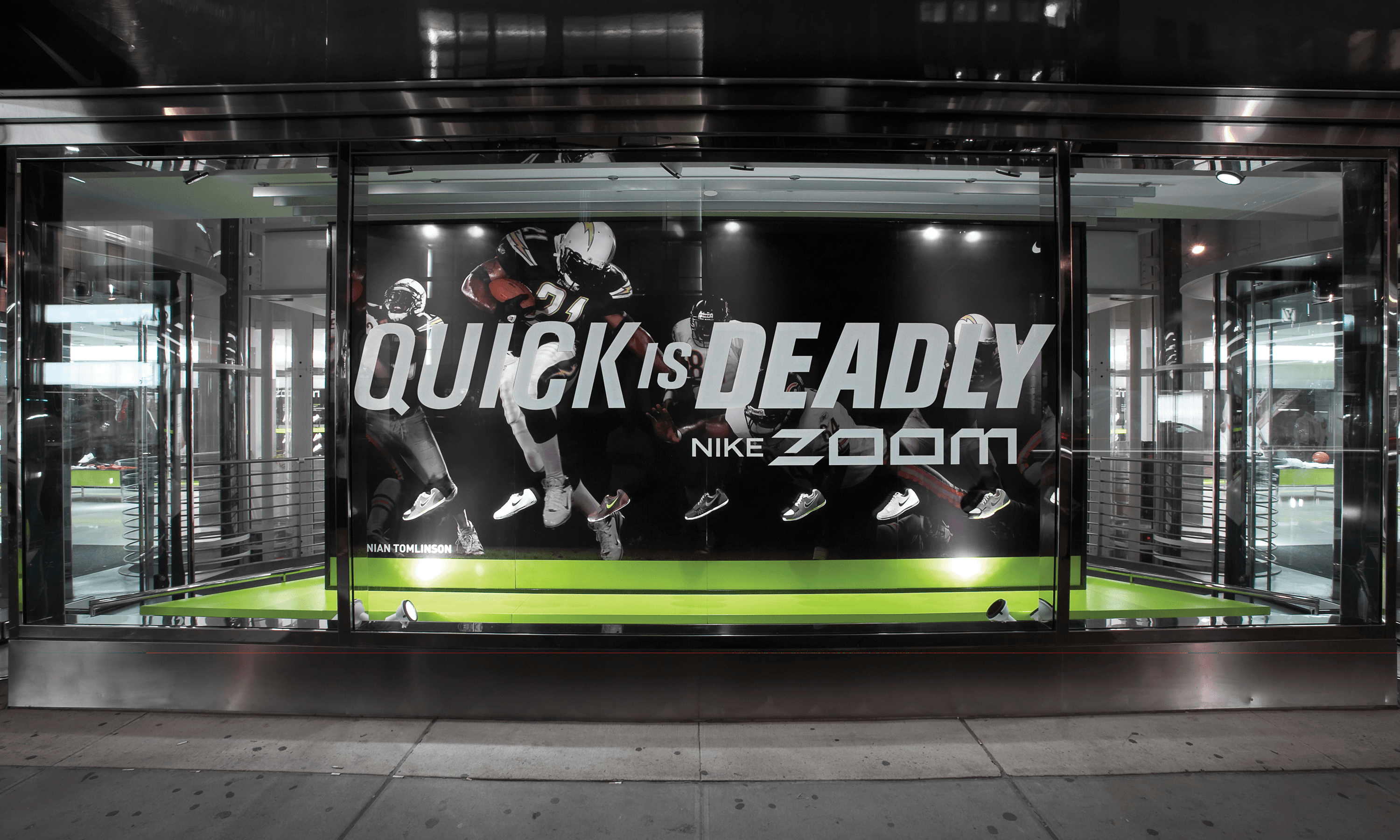 A street-level retail Nike Zoom window display at Niketown in New York City. The headline Quick Is Deadly is centered in the frame, with seven different shoes below in front of an image of football players playing football.