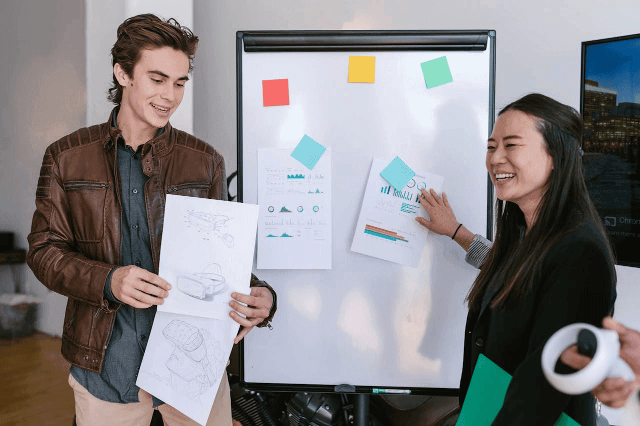 Two colleagues smiling while looking at charts and diagrams on a whiteboard in a collaborative setting.