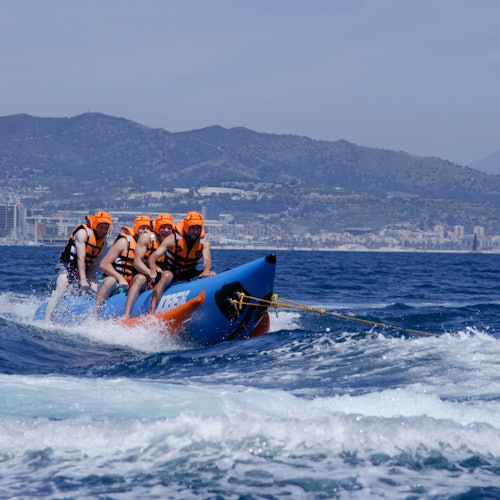 四个人戴着橙色头盔和救生衣，乘坐蓝色充气香蕉船在海上航行，背景是山峦。