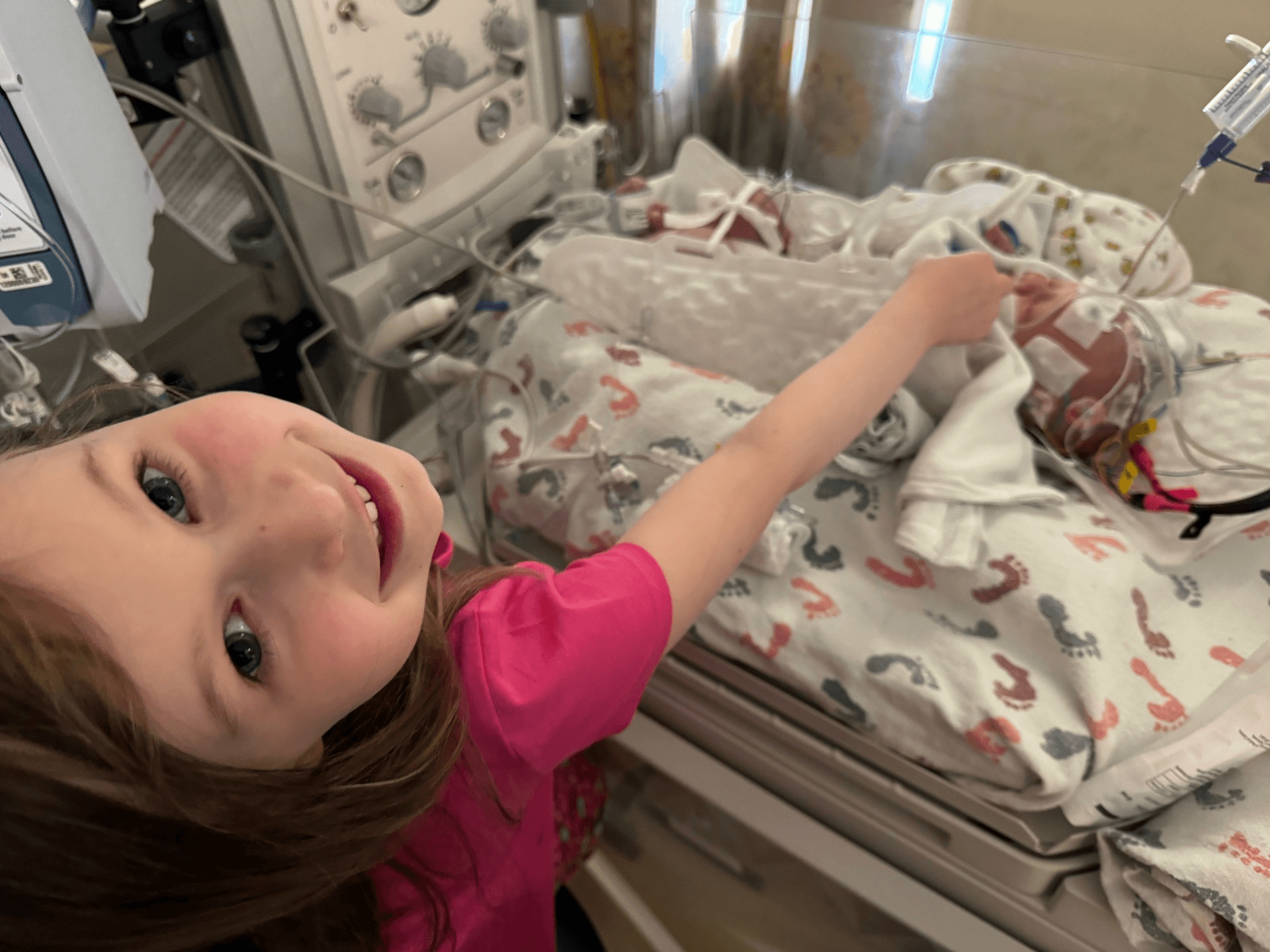 Young child gently touching a newborn baby in a hospital incubator, with medical monitors and tubing visible
