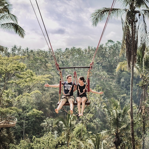 A man and woman are sitting on a swing suspended high above a lush, tropical forest with palm trees in the background.