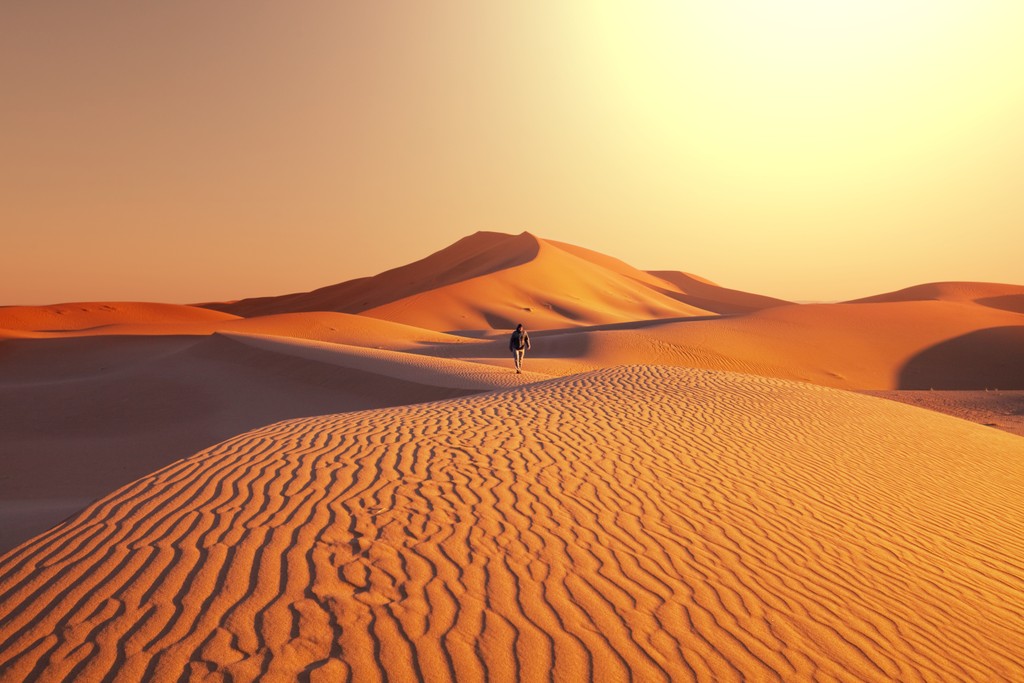 Solitary traveler walking across golden sand dunes at sunrise, with intricate wind-sculpted patterns and a warm, glowing horizon in the Namib Desert.