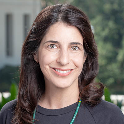 A light-skinned person with long, dark wavy hair smiles warmly at the camera. They are wearing a dark top and a green beaded necklace. The background shows blurred greenery and part of a white building.