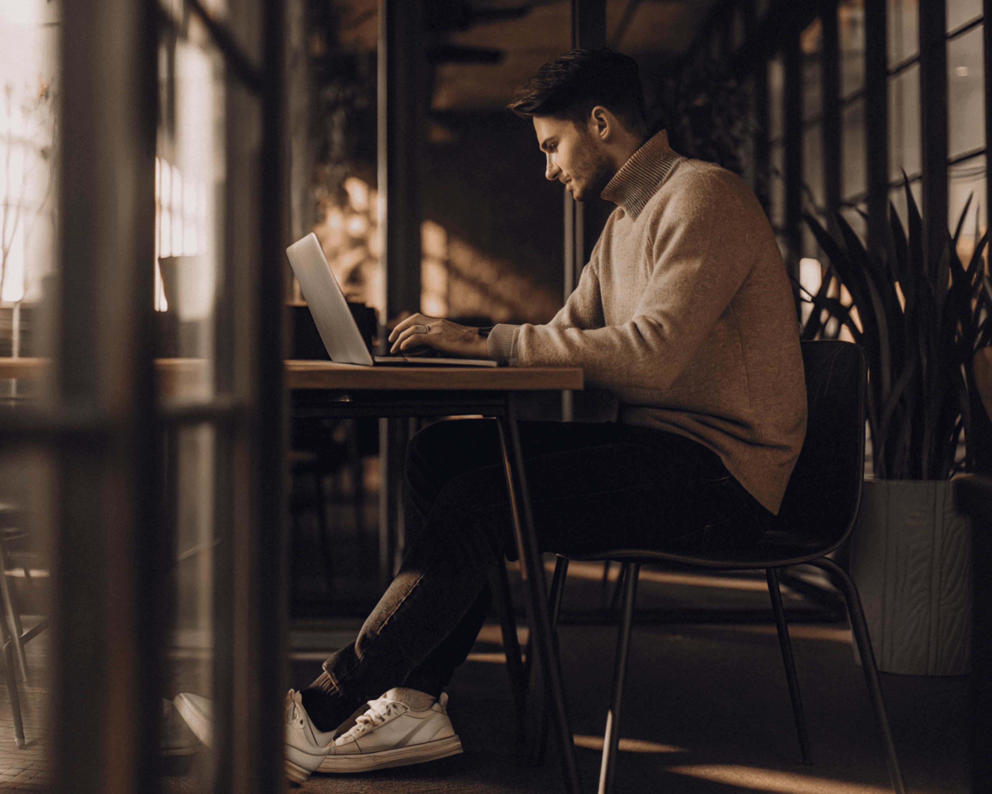 Man working on a laptop at a table, illuminated by natural light from a window. He wears a sweater, looking focused.