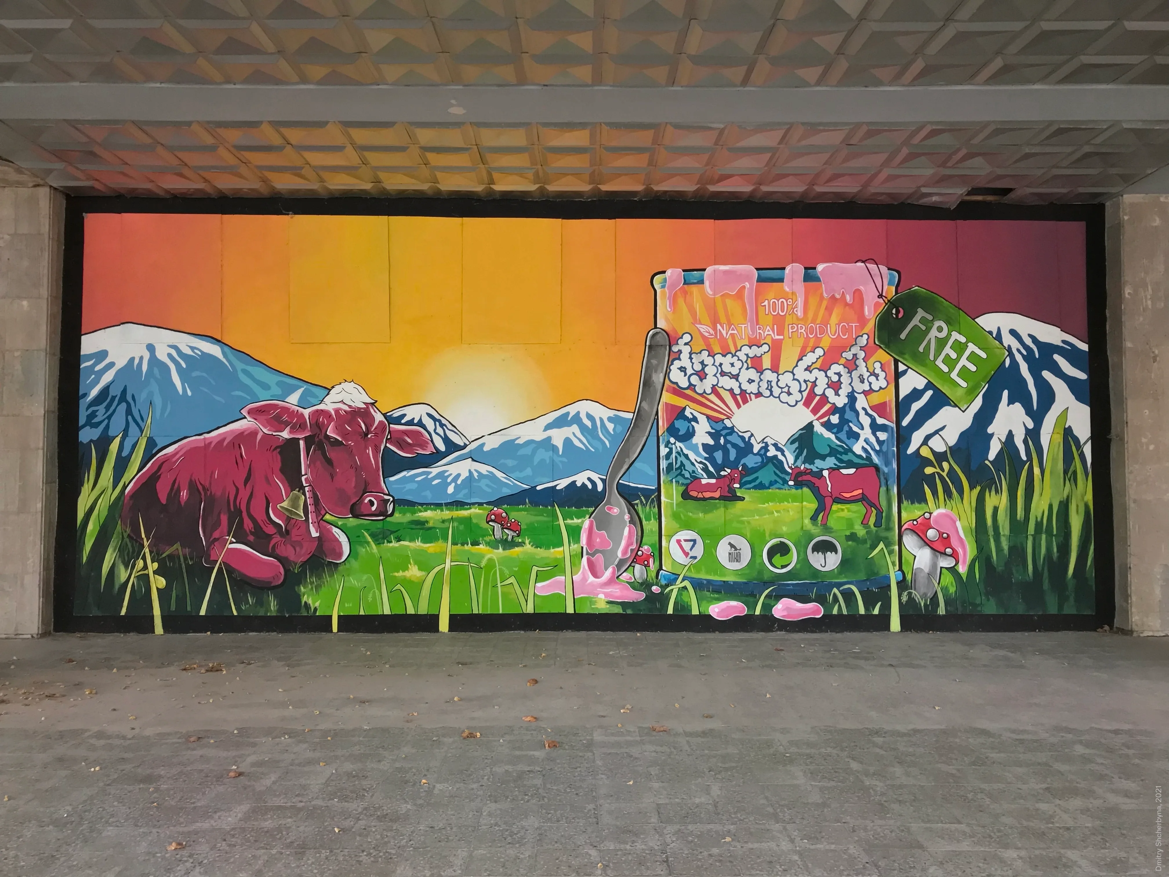 A mural depicting a pink cow resting in a green alpine meadow before snow-capped mountains and a sunset sky. Next to it sits a giant yogurt container labeled "100% Natural Product" in Georgian script, with a spoon, a "FREE" price tag, and mushrooms scattered around.