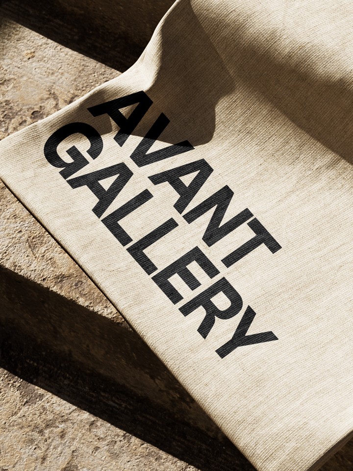 Beige canvas tote bag draped on a stone bench, printed diagonally in black with the stacked words “AVANT GALLERY”.