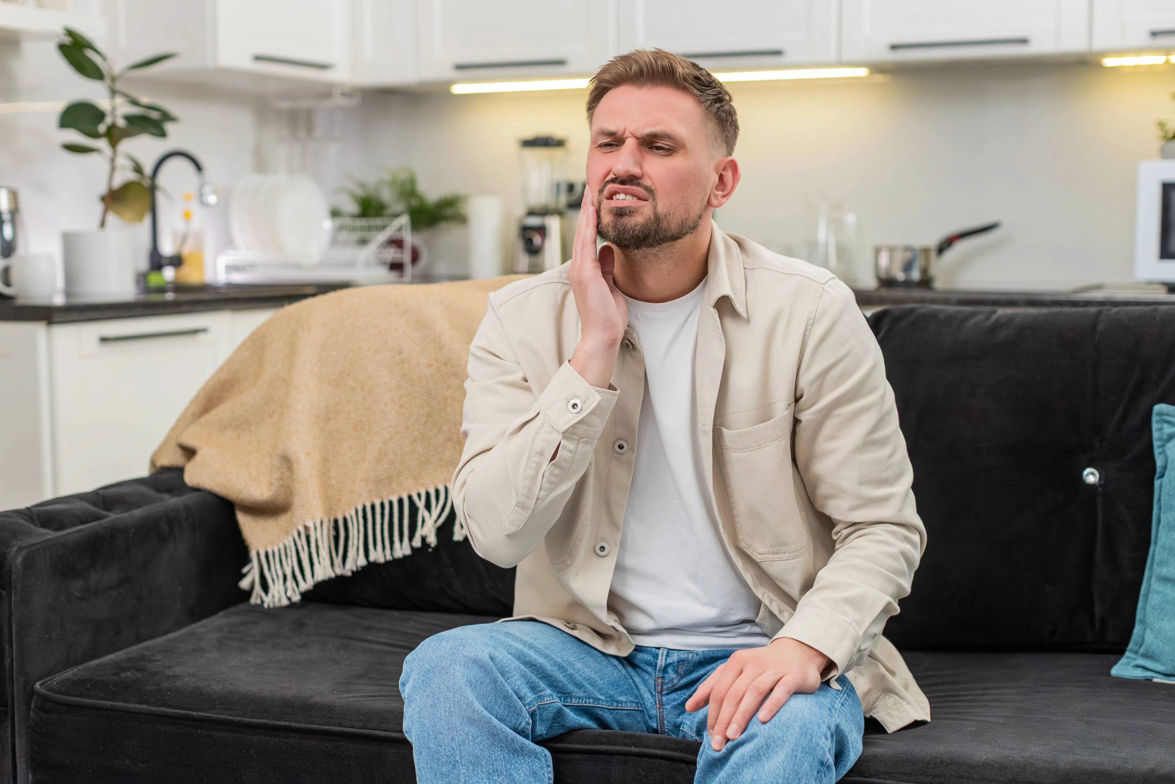 A man in casual attire sits on a black couch in a cozy living room, clutching his jaw in pain, suggesting a toothache. The space is warm and homey.