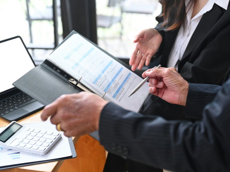 Two professionals engaged in a discussion over a document on a clipboard, with a laptop, cup of coffee, and eyeglasses on a wooden table, indicating a productive work meeting.