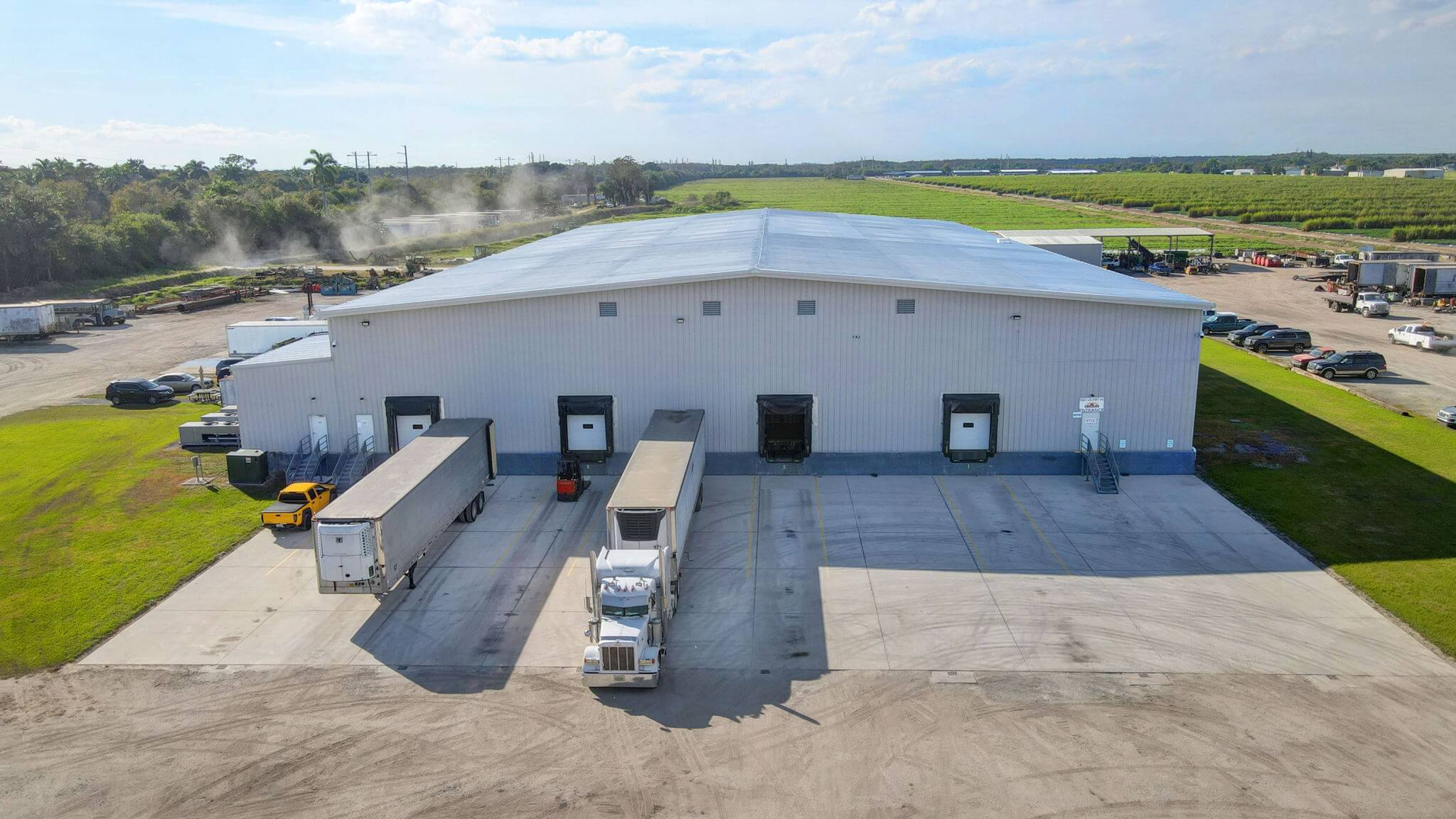 Completed white metal warehouse building with multiple loading docks and semi-trucks, featuring large concrete delivery area and metal roof system surrounded by green fields