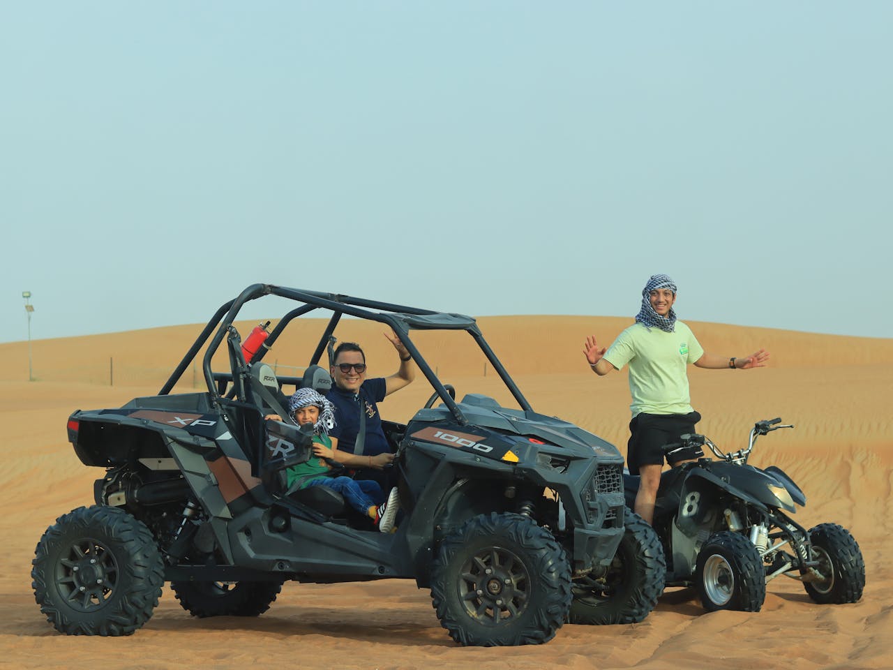 A father and his son are sitting in a dune buggy while another person is on a quad bike in the desert.