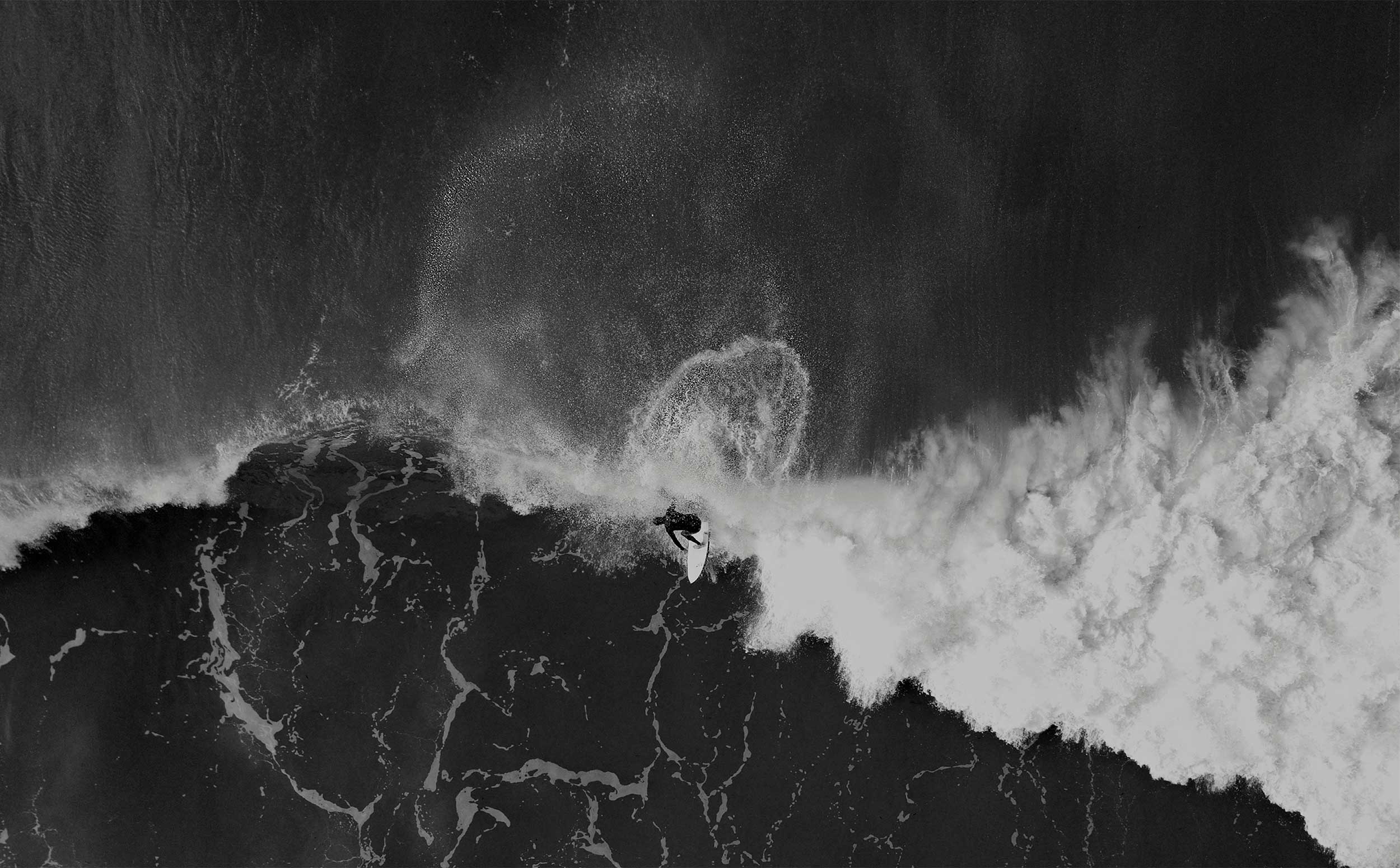A black and white aerial photo of a surfer riding a wave. The image shows the surfer carving through the water, creating a dynamic spray of water around them.