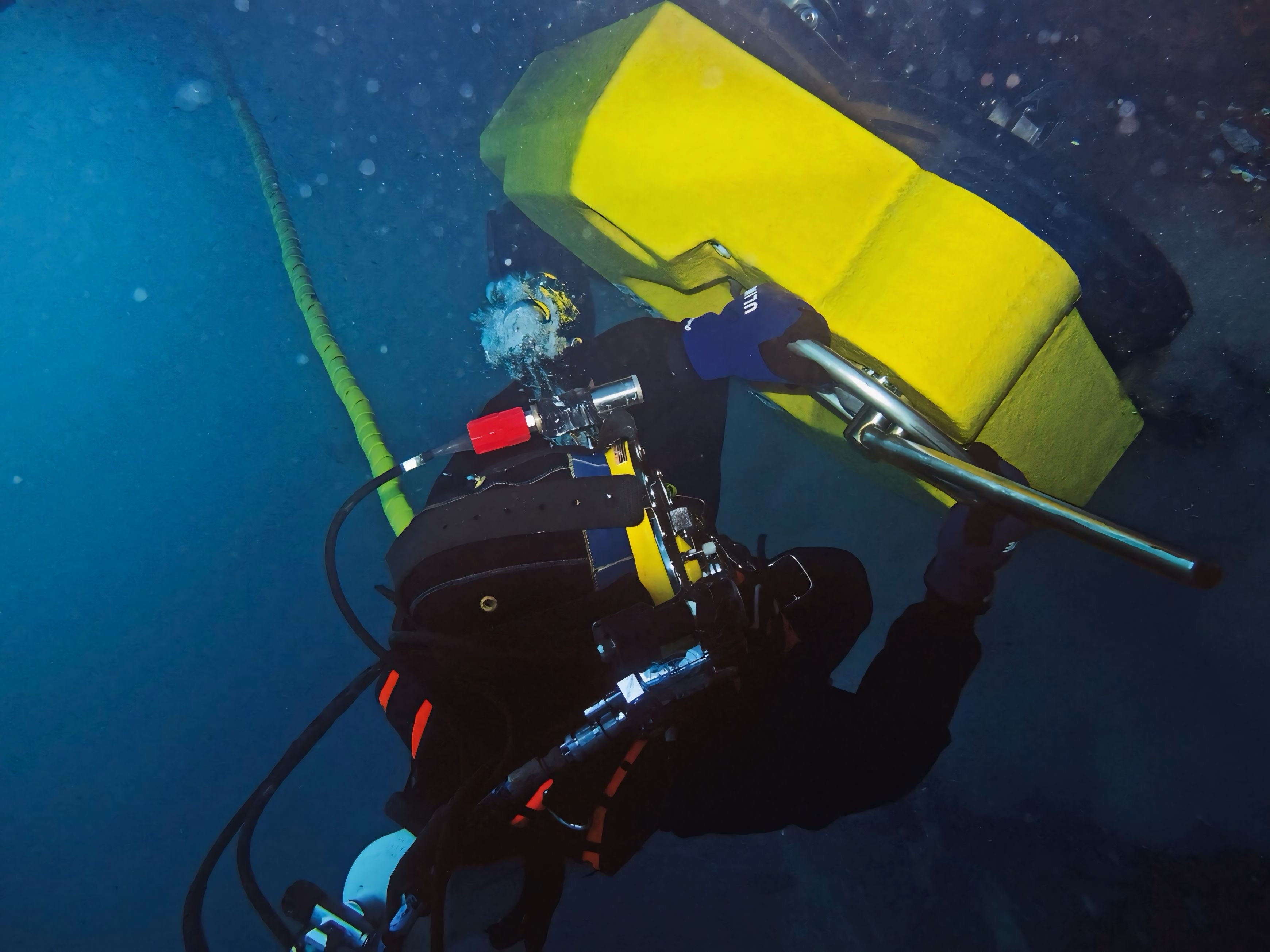 Seachios® Marine Services commercial diver operating an advanced capture-type underwater hull cleaning system, firmly handling the yellow cleaning unit and umbilical hose against a vessel’s hull to perform eco-compliant UWHC and underwater inspection services.