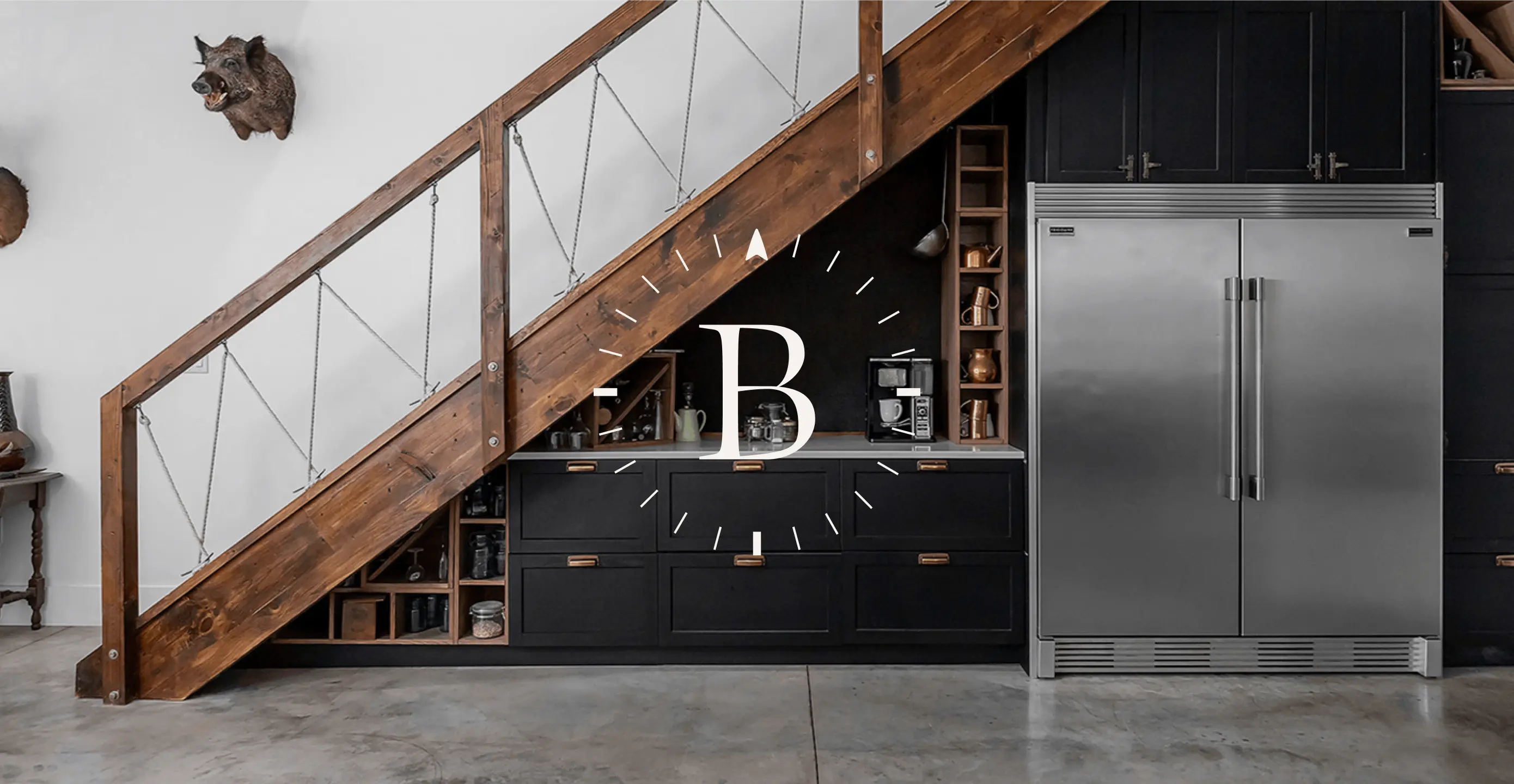 Under-stair kitchen with black cabinets, a large fridge, and a rustic wooden staircase.