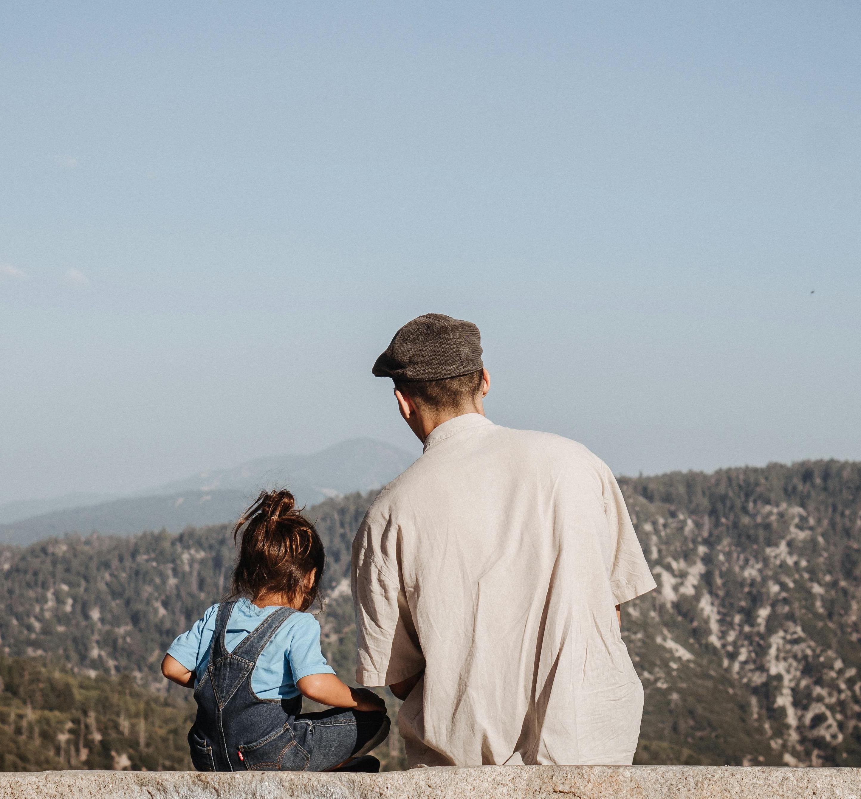 A man in a cap and a young child sit on a stone ledge, overlooking a vast mountain landscape under a clear blue sky.
