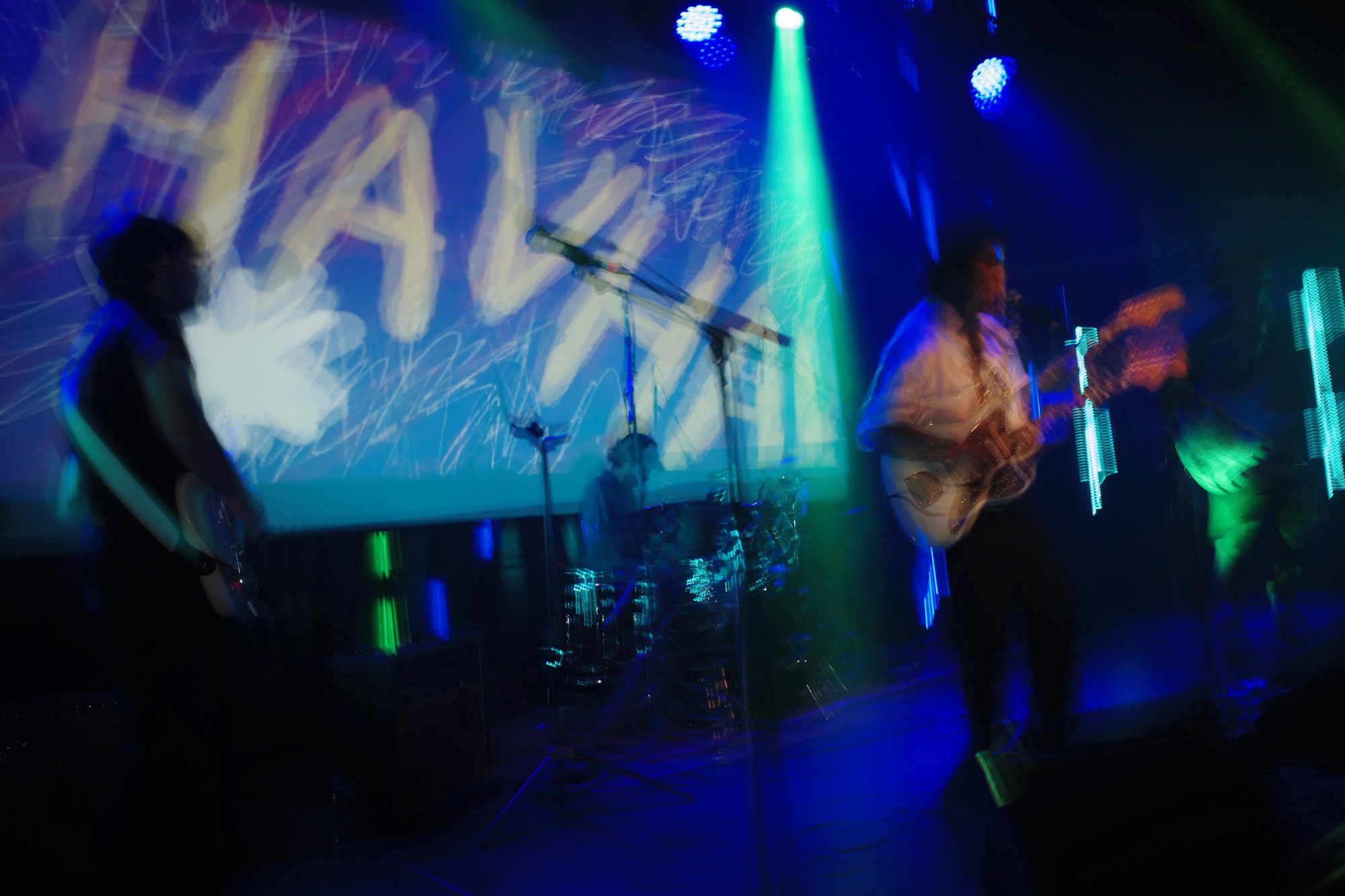 A vibrant live music performance with a band playing on stage, featuring colorful stage lights and a large screen displaying abstract graphics in the background.