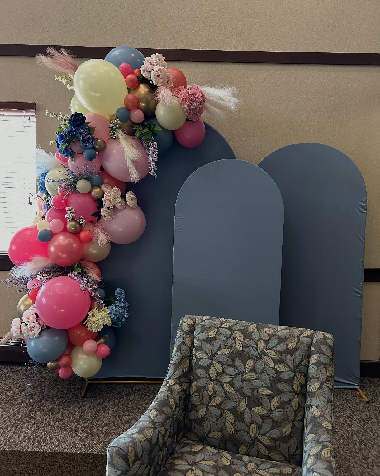 Balloon garland with pink, peach, and gold balloons arranged around blue backdrop panels and a patterned chair by Be Bold Balloons