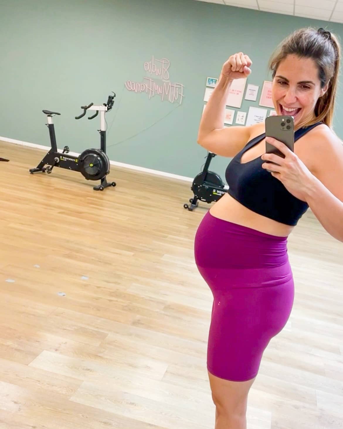 Selfie miroir de Caroline Rinaudo, coach FitMum France enceinte, souriante et montrant son muscle dans sa salle de sport, portant une tenue de sport noire et violette.
