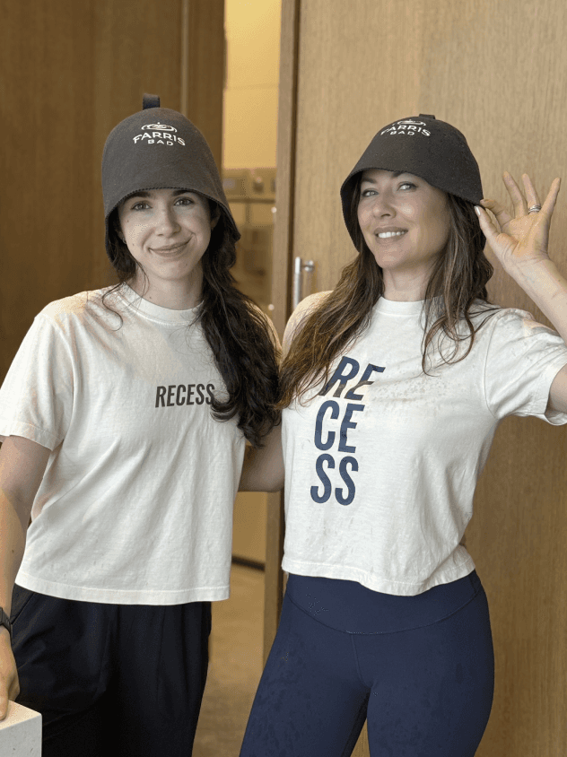 A person relaxes in a round, black outdoor hot tub on a rooftop, surrounded by glass railings with a view of distant trees and a modern apartment building under a cloudy sky.