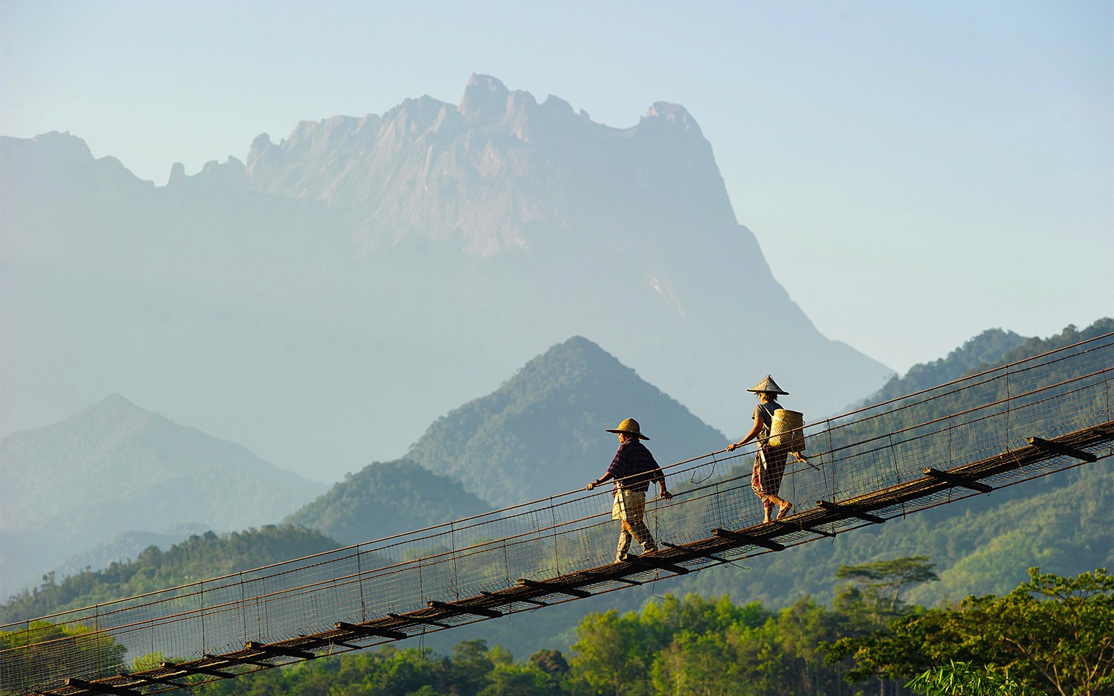 Hikers crossing a suspension bridge with Mount Kinabalu in the background.