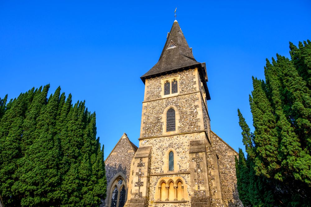 A smiling man with curly hair is sitting at a desk, looking at the camera, with a laptop and a lamp in the background.
