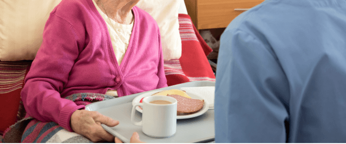 Caregiver serving meal to elderly person in bed.