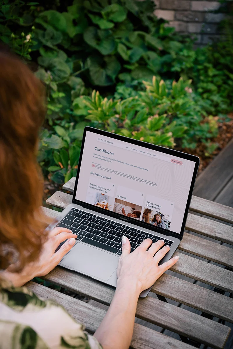 A woman sits at an outdoor table using a laptop to view the Lift and Revive website, looking at a conditions page with information on bladder control and pelvic health concerns.