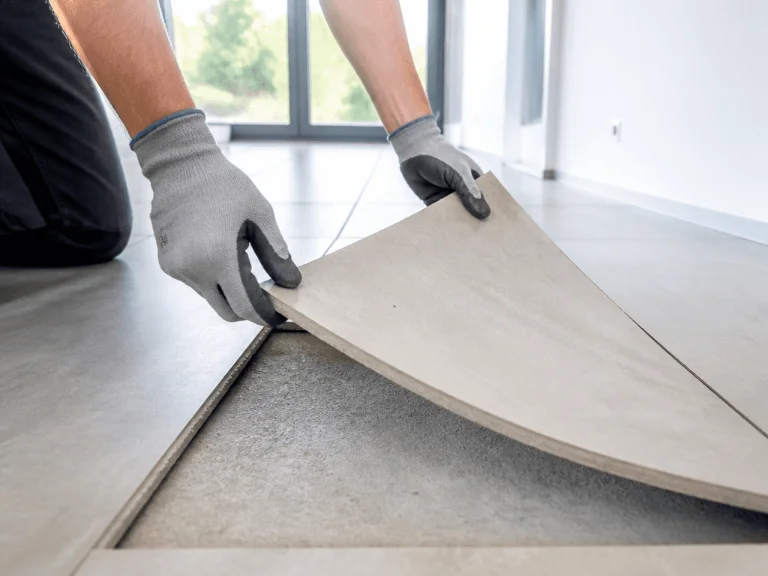 Eine Person auf den Knien, die mit grauen Arbeitshandschuhen eine quadratische, hellgraue Bodenfliese vom Untergrund anhebt, um den darunter liegenden Estrich freizulegen.