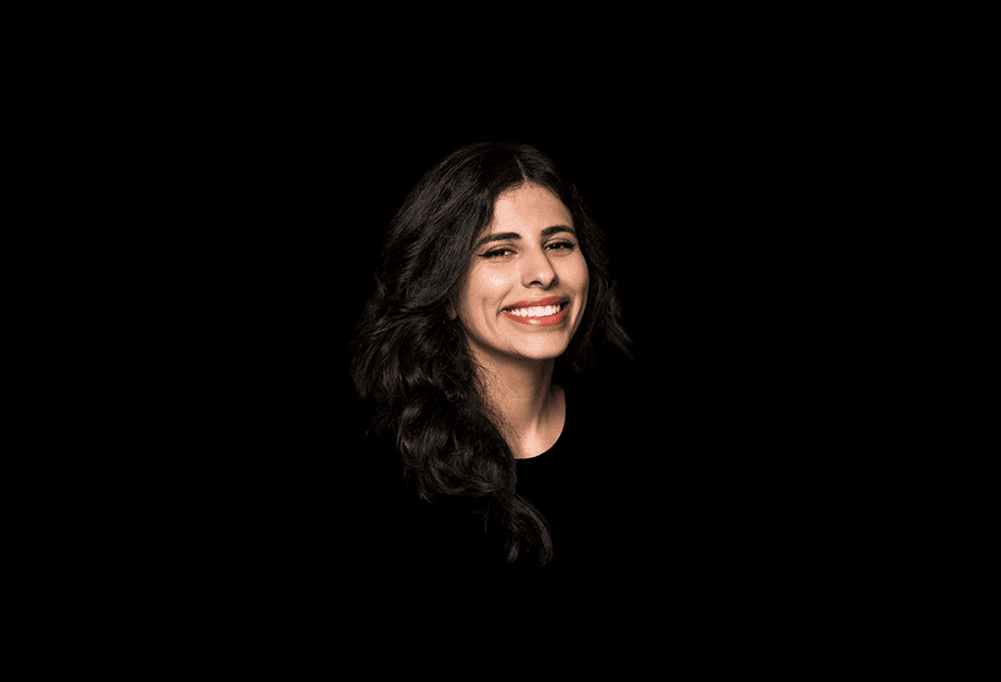 A woman with long, dark hair is shown against a solid black background. She is smiling and looking directly at the camera. Her hair falls around her shoulders. She is wearing a dark-colored top. The lighting is focused on her face.