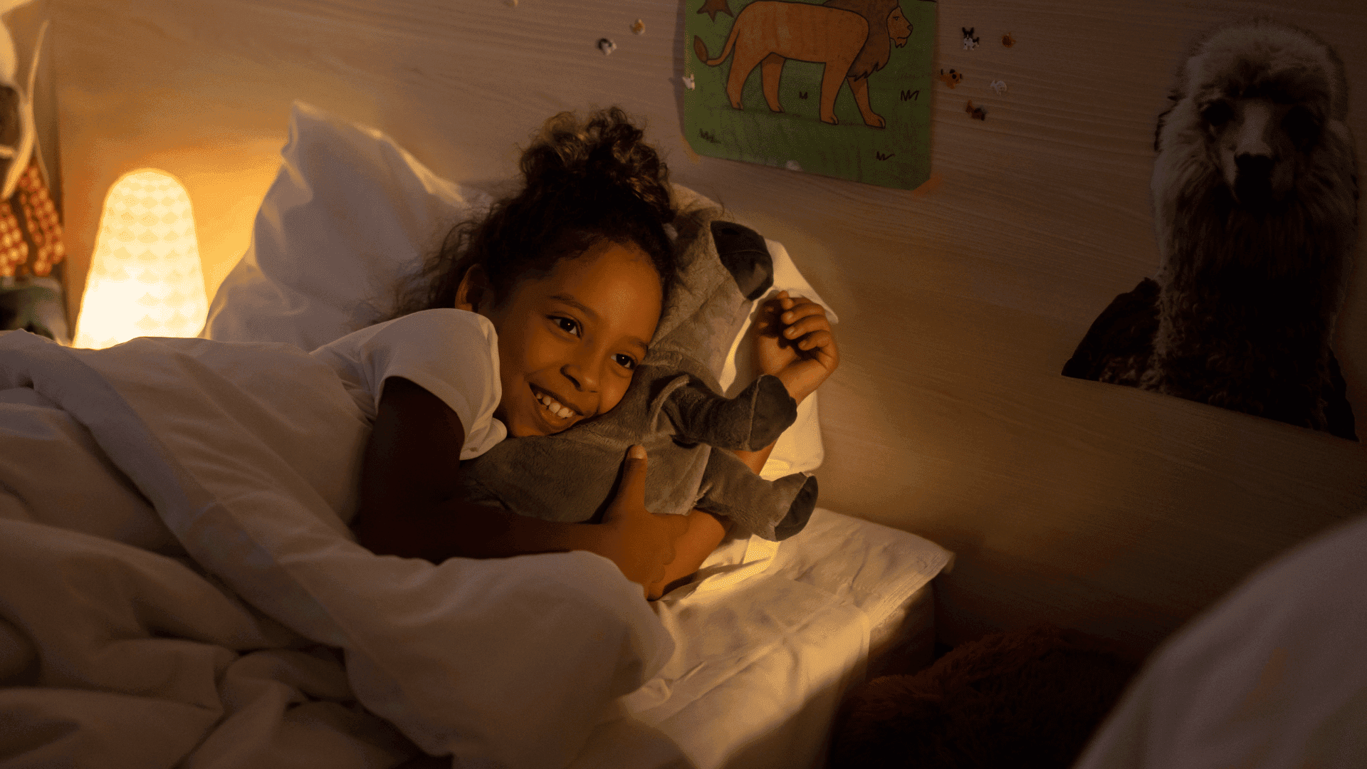 A young girl tucked in bed, smiling and looking relaxed, with a night light beside her.