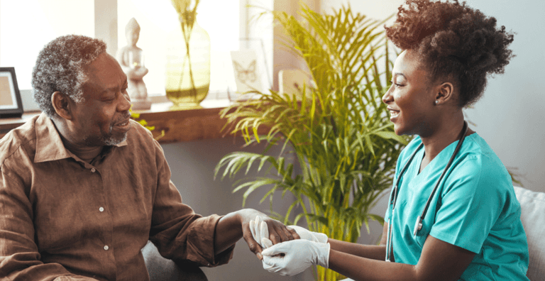Caregiver in scrubs gently holding an older man’s hand during a home visit.