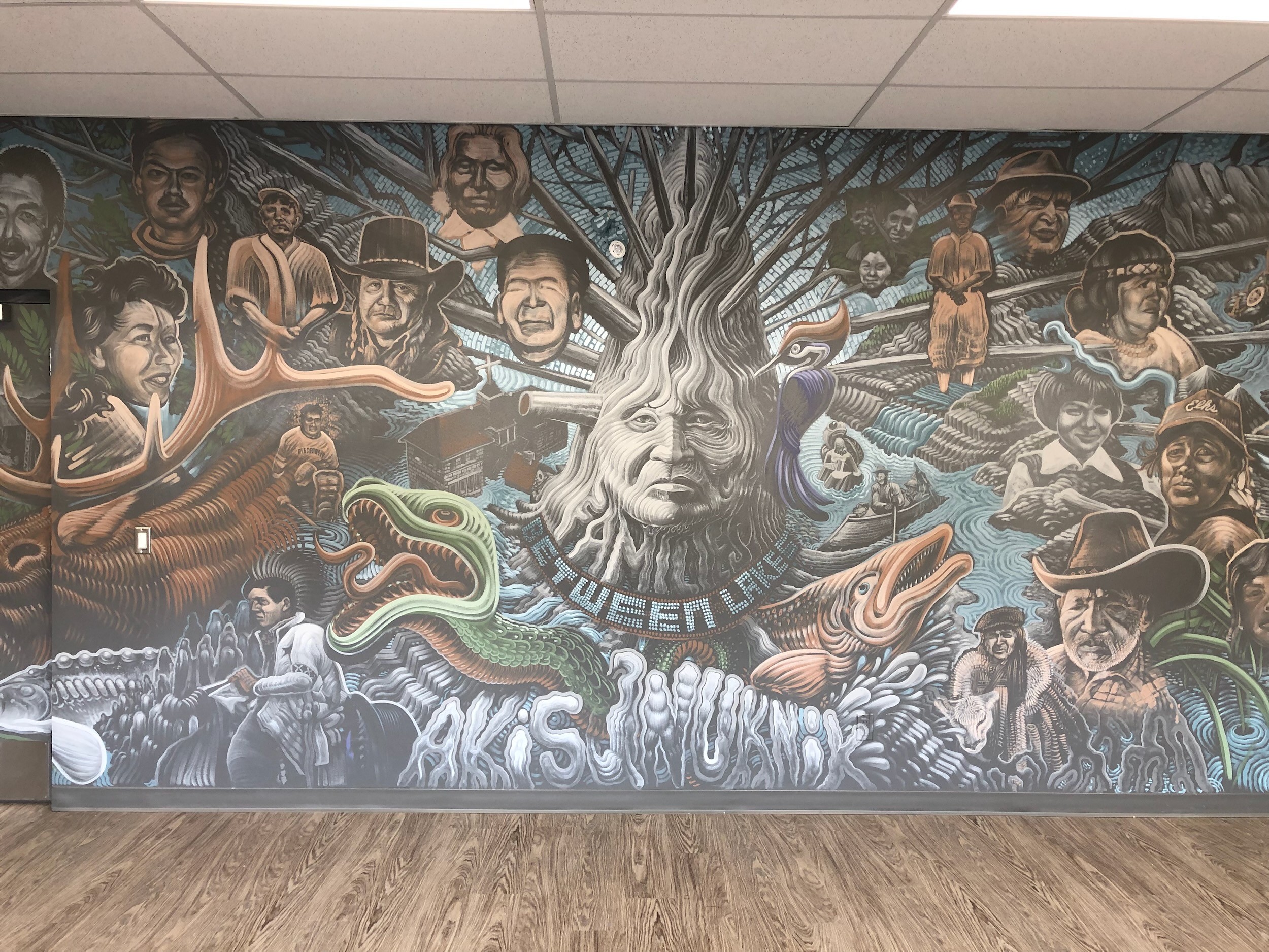 A wall of painted portraits gathered around a central face rooted like a tree, a serpent and salmon at its foot.