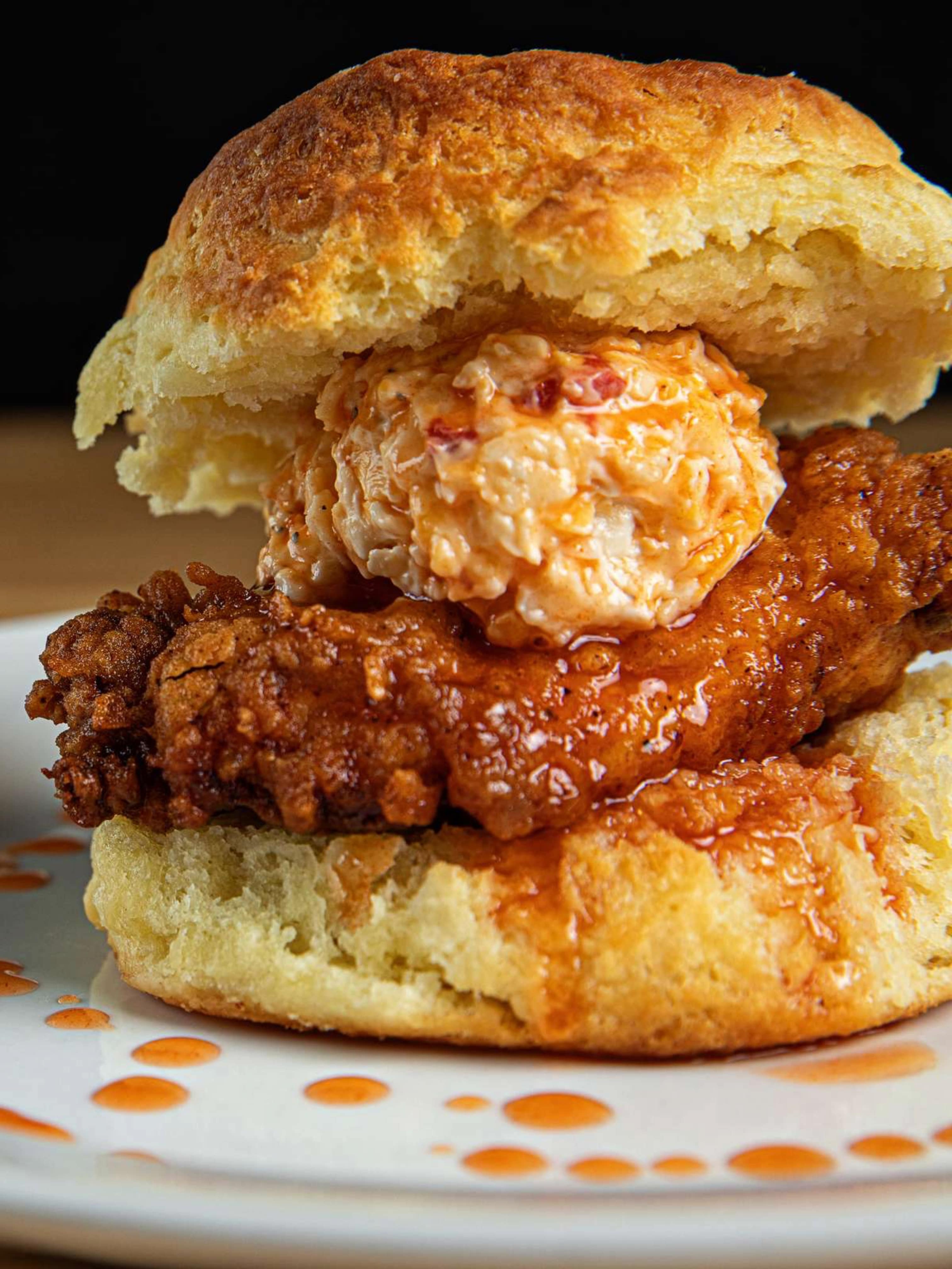 A mouthwatering chicken biscuit sandwich from Vicious Biscuit, featuring a crispy fried chicken breast topped with pimento cheese and drizzled with hot honey, served on a fluffy biscuit. The dish is plated on a white plate with a decorative drizzle of sauce.