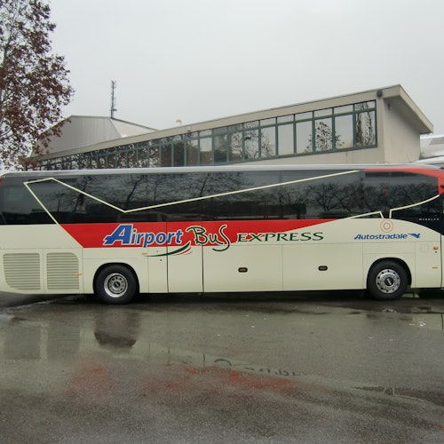 一輛「機場巴士快線」停在一棟建築旁邊，路面潮濕，天色陰雲。