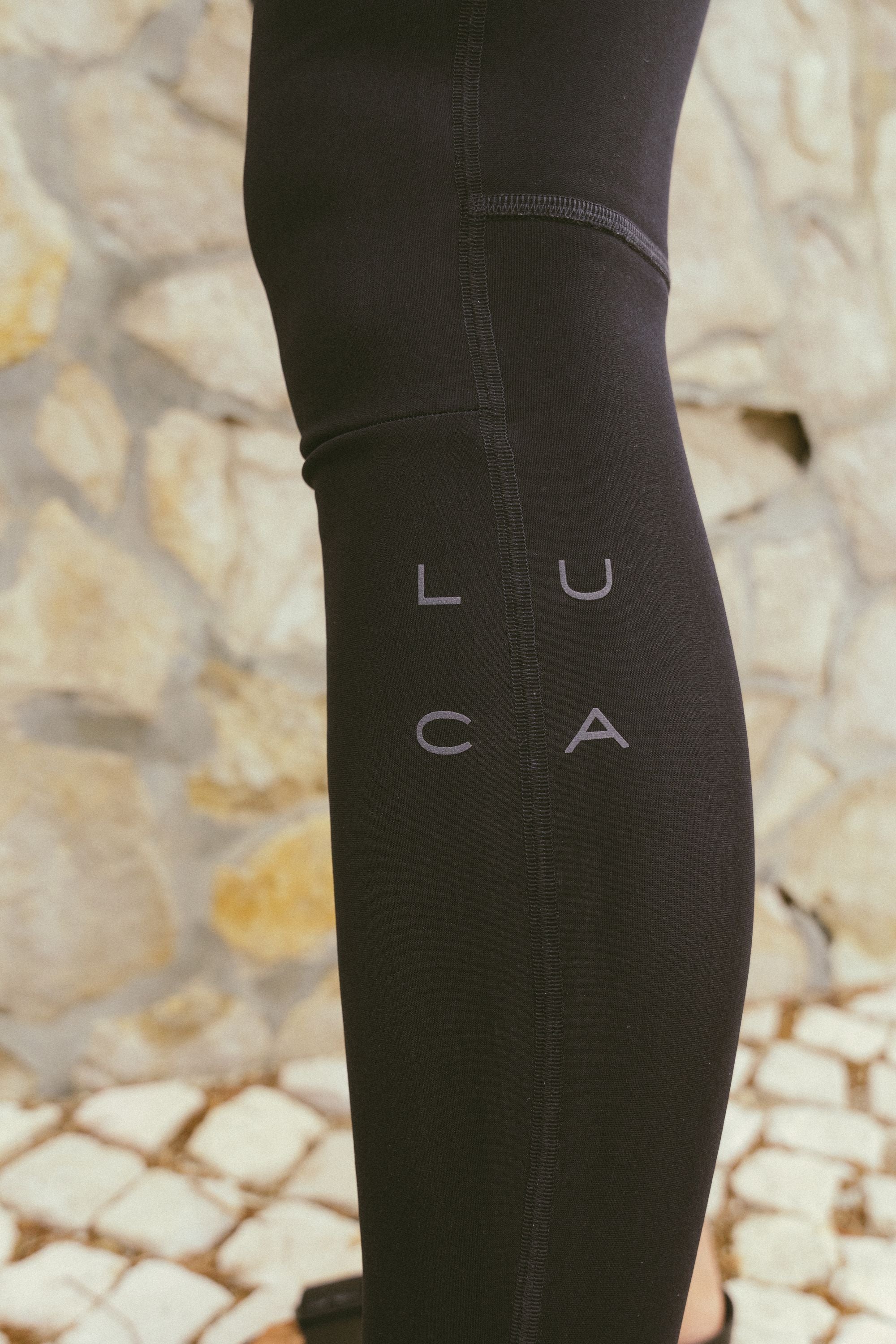 Extreme-close-up-of-a-person's-leg-wearing-solid-black-tight-fitting- Stride-Bib-Tight-The-black-fabric- features-a-subtle-matte-gray-LUCA- logo-printed-vertically-on-the-calf- arranged-in-two-rows-of-two-letters-The-background-shows- blurred-light-colored-stone-paving- and-a-textured-natural-stone-wall