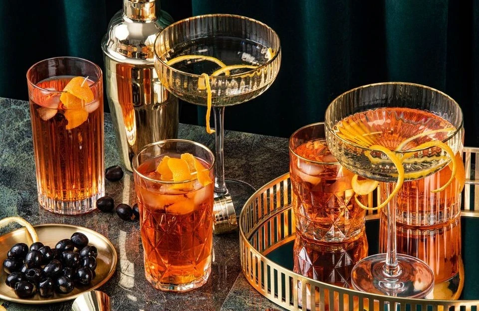 Detailed close-up photo of premium cocktails, including an espresso martini and two highball drinks, served on a textured tray with a bowl of black olives, suggesting luxury.