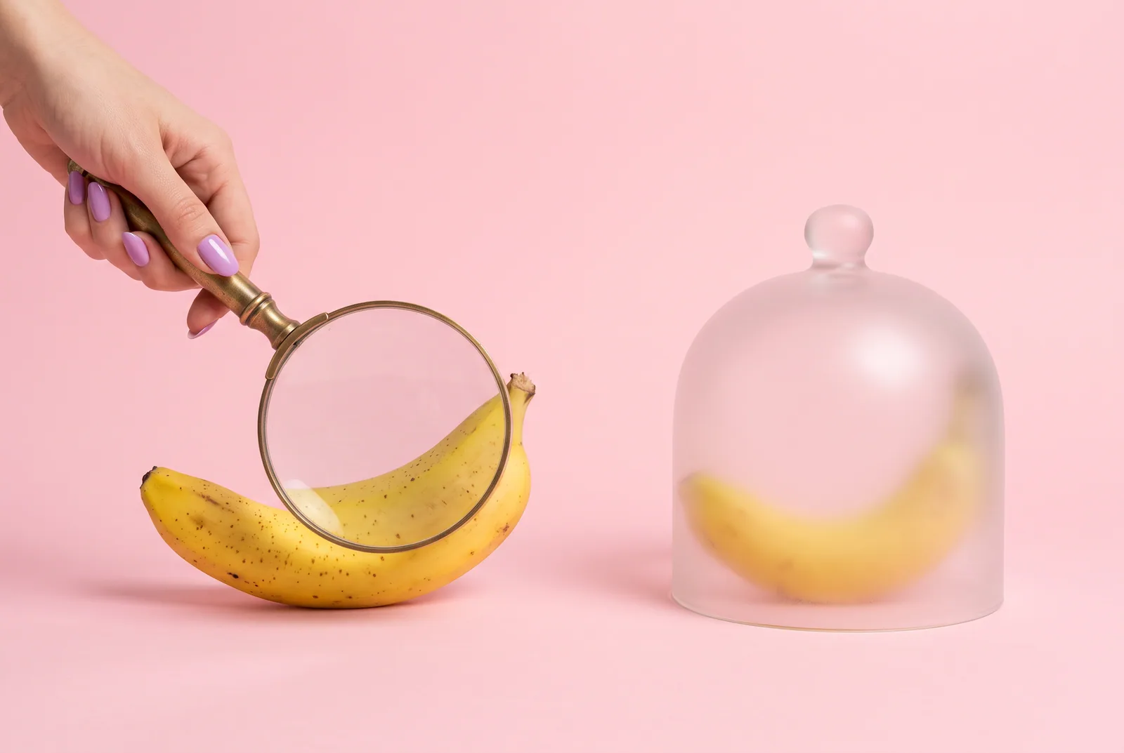 Porn accountability software concept: a hand holding a magnifying glass over a banana while a second banana rests under a frosted glass cloche