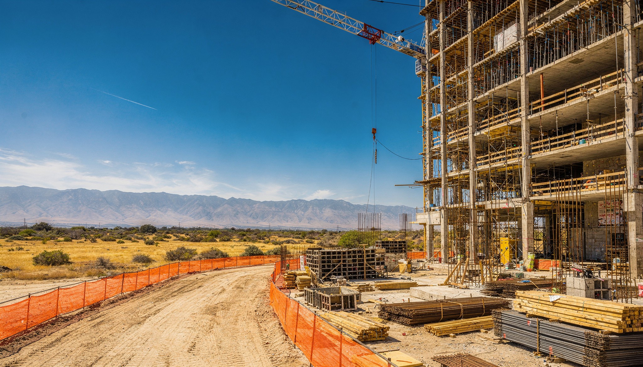 A construction site on flat, arid terrain under an overcast sky. Workers wearing reflective vests are working in a trench protected by an orange fence, while an orange-and-white excavator moves earth in the background, near a large, cylindrical elevated water tank.