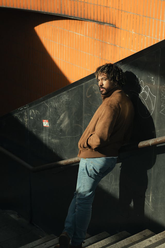 A man in a leather jacket and light-wash jeans sits in an alley