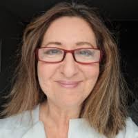 A young person with curly hair smiles at the camera while sitting in front of a laptop against a dark background.