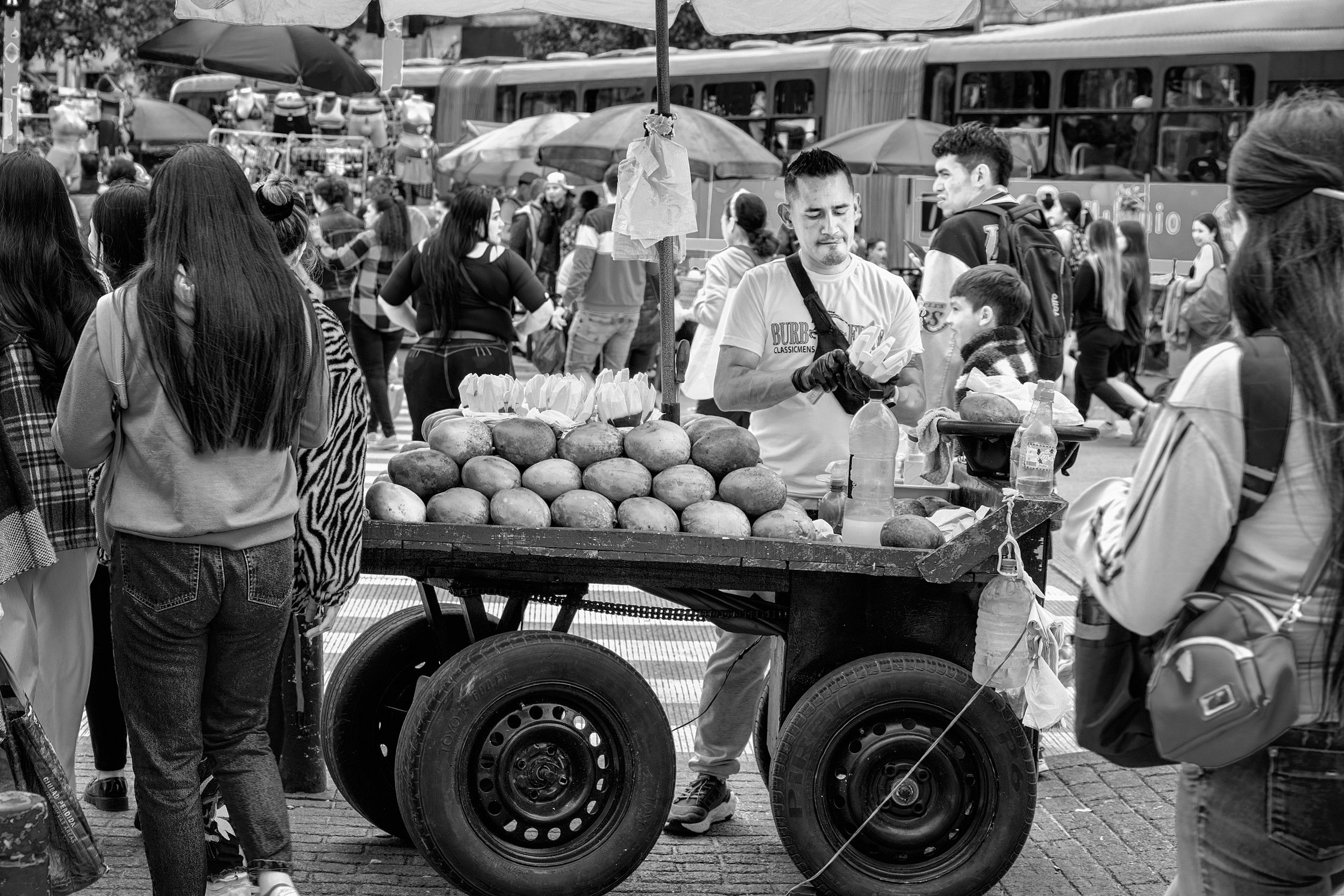 A vendor stands behind a cart stacked with ripe mangoes, preparing servings for a crowd of customers at a busy intersection in Bogotá.