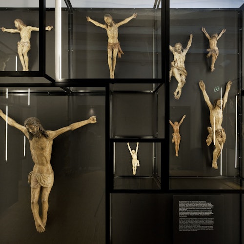 Nine crucifix sculptures of Jesus displayed in a glass case, each varying in size and pose.